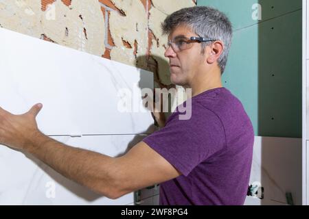Side portrait of a tiler wearing protective glasses placing a large format tile piece on a brick wall in a bathroom renovation. Home renovation concep Stock Photo