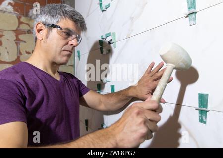 Portrait of a tiler wearing protective glasses fixing a piece of tile with tapping wedges with a rubber mallet in a bathroom renovation. Home renovati Stock Photo