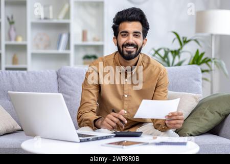 Close-up portrait of a young Indian man working remotely from home, sitting on a sofa, holding documents and using a calculator, smiling at the camera. Stock Photo