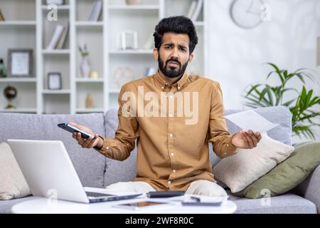 Portrait of upset and charming young Indian man sitting on sofa at home, looking at camera and holding bills and phone. Stock Photo