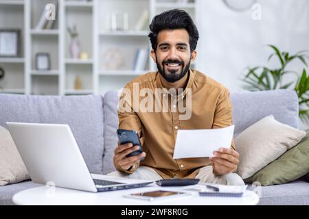 Portrait of a young Indian man sitting at home on the couch, holding a phone and documents, bills. Looking and smiling at the camera. Stock Photo