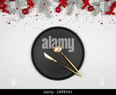 Christmas composition, black plate and gold cutlery, fir tree branches, red berries, baubles on white background. Stock Photo