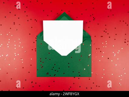 Top view of green envelope, white card and stars confetti on red background. Christmas, New Year composition. Stock Photo