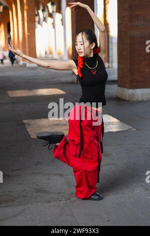 An urban dancer in striking red trousers performs an elegant pose inspired by oriental dance on a city walkway. Stock Photo