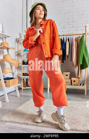 confident asian designer in orange clothes and panama hat posing in modern personal atelier Stock Photo