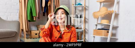 joyful asian stylist posing in orange attire and panama hat in atelier, fashion business, banner Stock Photo