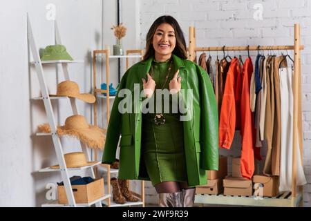 joyful asian personal stylist posing in trendy clothes and green leather jacket in fashion studio Stock Photo