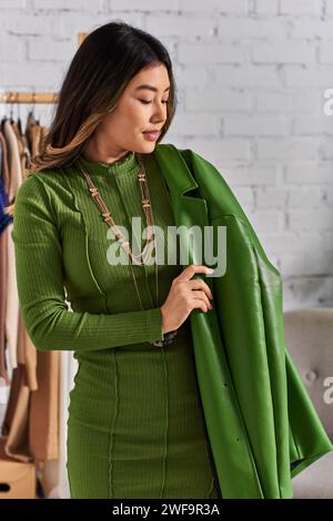 trendy asian dressmaker posing in green leather jacket in private fashion atelier, small business Stock Photo