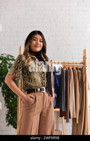 joyful asian stylist in fashionable casual attire posing with hands in pockets in personal atelier Stock Photo