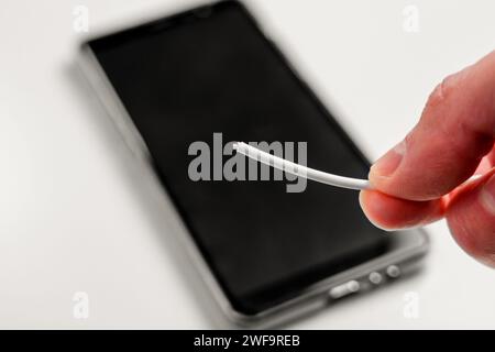 Hand holding a broken torn charger cable with a black smartphone on a white background. Energy connect problem concept Stock Photo