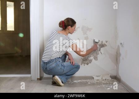 Saltpeter on the wall problem. Woman is using a scraper to scrape and remove all loose paint and plaster that is in poor condition, until a firm surfa Stock Photo