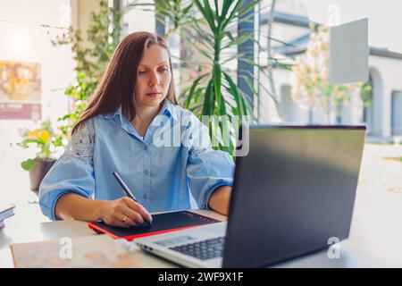 Young female teacher getting ready for lesson working in office using laptop computer. Woman writing notes on tablet Stock Photo