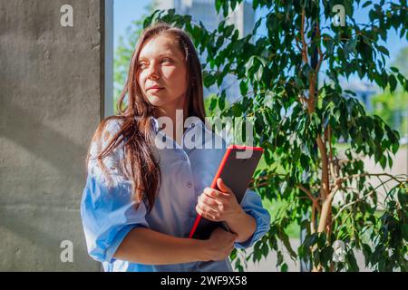 Happy confident office IT worker holding tablet standing by window. Caucasian young woman looking at camera. Business concept. Space Stock Photo