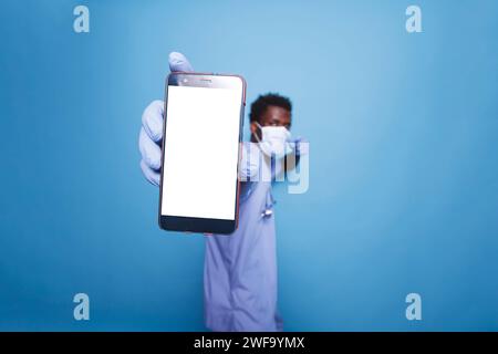 Nurse of African American ethnicity in scrubs and face mask holds a smartphone displaying blank chromakey mockup template. Black man grasps mobile device with white screen close to camera. Stock Photo