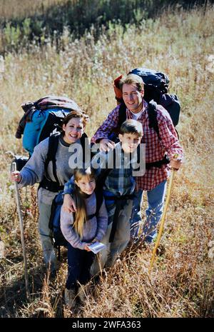 A Portrait of a happy family with two children and backpacks is smiling at the camera while hiking in a grassy field on a sunny autumn day. Stock Photo