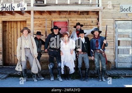 A group of seven people stands in a line, posing in period-specific costumes representative of the American Wild West era, in front of a rustic saloon Stock Photo