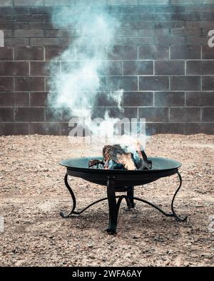 Smoke rising from a burning fire in a backyard fire pit with wooden logs, brick wall in the background. Stock Photo