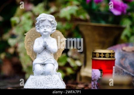 a small white angel figure kneels praying on a grave in front of a grave candle and gravestone as well as flowers in the blurred background Stock Photo