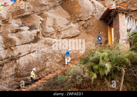 Bhutanese man wearing traditonal gho and female tourists climb the steep stairs on the rocky path to Tiger's Nest monastery, ready to pass through a d Stock Photo