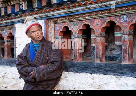 An animated old man stands in front of prayer wheels at an ancient Buddhist temple; Ogyen Choling village, Bumthang Valley, Bhutan, Asia Stock Photo