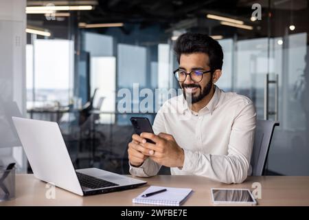 A young Indian man is sitting in an office at a desk and smilingly using a mobile phone. Stock Photo