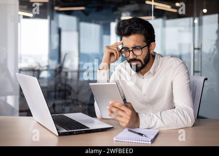 Thoughtful and serious young Indian male businessman sitting in the office at the table and using a tablet. Stock Photo