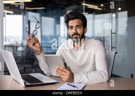 Portrait of a serious Indian young man businessman sitting in the office at the desk and looking at the camera, holding glasses and working with a tablet. Stock Photo