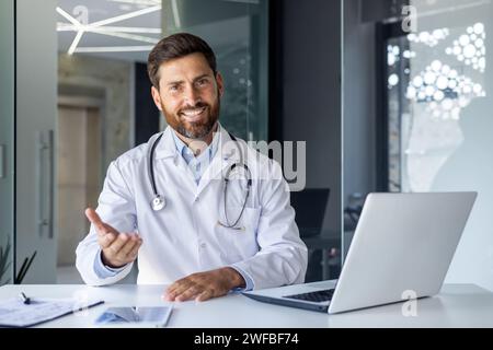 Portrait of a smiling young male doctor talking and consulting on a video call. He is sitting at the table in the office of the clinic, gesturing with his hand in front of the camera. Stock Photo