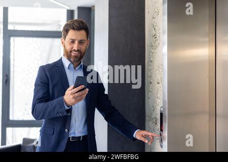 A smiling young male businessman is standing in an office space, calling an elevator and using a mobile phone. Stock Photo