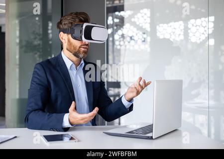 Businessman in suit using VR headset at desk in modern office, gesturing with hands, exploring virtual reality. Stock Photo