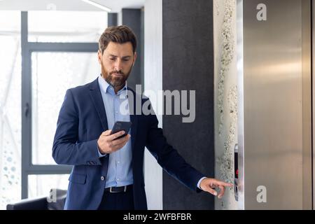 Focused businessman in a suit using smartphone while waiting for an elevator in a modern office building interior. Stock Photo