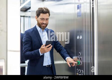 A young smiling businessman and lawyer is standing in the elevator of an office building and pressing the wrong button while using a mobile phone. Stock Photo