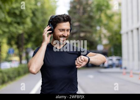 Close-up photo of a smiling young male athlete standing outside wearing headphones and looking contentedly at a smart watch on his wrist. Stock Photo