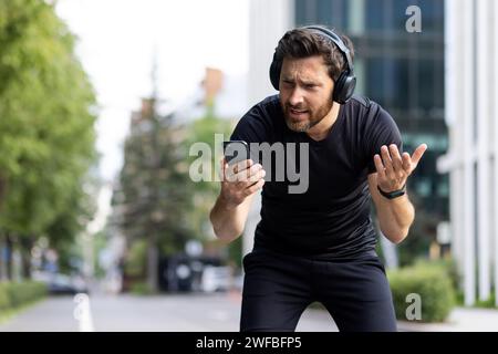A young male sportsman in headphones stands bent over tired on the street, resting. He looks disappointedly at the phone screen, spreads his hands. Stock Photo