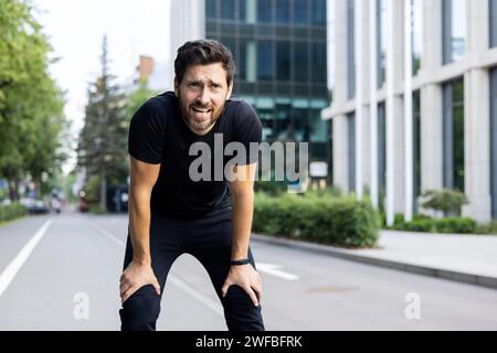 Portrait of tired young man standing bent over on street road after jogging and breathing hard, resting after marathon and training. Stock Photo
