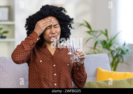 Worried young African American woman is at home, holding a glass of water and medicine in her hands, holding her head in pain, not feeling well. Stock Photo