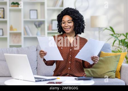 African American young businesswoman working remotely from home. He sits at home on the sofa and looks over documents, agreements, bills. Stock Photo