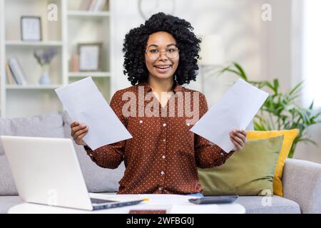 Portrait of a smiling African-American woman sitting at home on the couch at the table with a laptop, holding documents and papers in her hands, working remotely. Stock Photo
