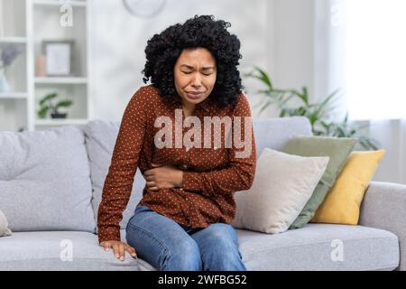 African american young woman sitting on sofa at home, closing eyes from pain and holding hand on stomach, feeling bad. Stock Photo