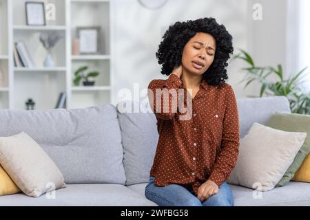 African American young woman sitting at home on sofa tired, closing eyes resting and doing neck massage, feeling pain and tension. Stock Photo