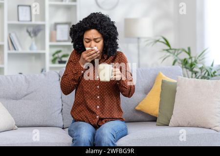 African american young woman wiping nose with tissue from cold, sitting on sofa at home sick and holding cup in hands. Stock Photo