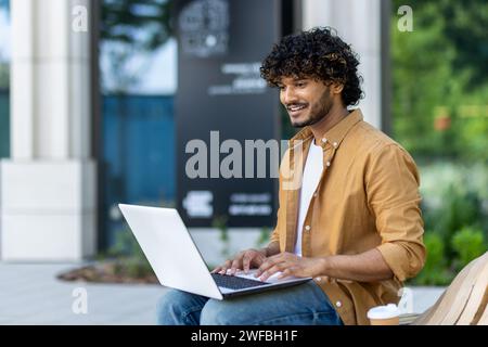 Indian young man sitting on the street with a laptop and texting, talking on a video call with a smile, studying, working. Stock Photo