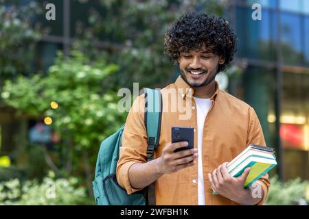 Indian smiling young male student standing outside with backpack and books and using mobile phone. Stock Photo