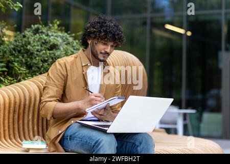 Serious young Muslim man sitting outside on a bench wearing headphones and using a laptop, writing in a notebook, listening and talking on a video call. Stock Photo