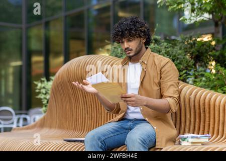 Shocked young Indian male student sitting on bench near office center and reading documents and letter, raising hands in frustration, worried about news received. Stock Photo