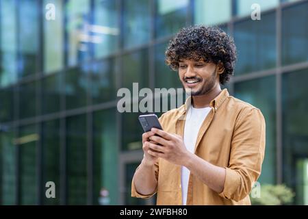A smiling young Indian man is standing on the street near an office center and using the phone, talking on a video call. Stock Photo