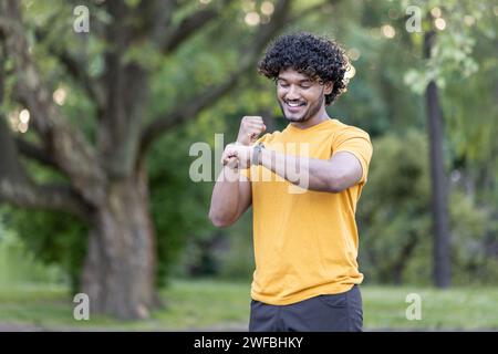 Smiling Indian young man running and doing sports. Standing in the park and looking at the smartwatch on his hand, he is happy with his success. Stock Photo