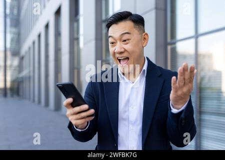 Happy Asian businessman standing outside in a suit and surprised and happy looking at the screen of his mobile phone. Stock Photo