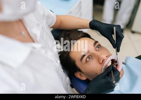 Top view of shocked male patient sitting in stomatology seat with open mouth during unrecognizable female dentist examining teeth, with dental tool in Stock Photo
