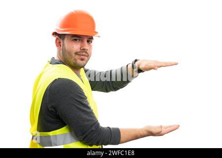 Constructor man with friendly smile wearing orange hardhat and fluorescent vest presenting small object copyspace between palms isolated on white back Stock Photo
